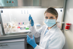 Female scientist wearing gown, mask and gloves, looking at the camera holding a beaker and pipet.