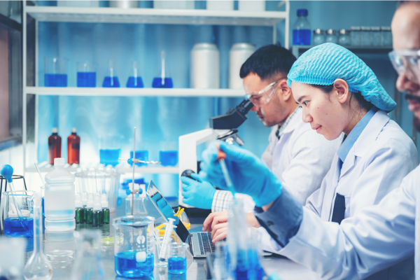 Two scientists, one female, one male, working in a lab. The make is looking into a microscope and the female is using a pipet.